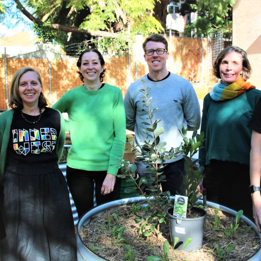 Four people smiling at the viewer standing behind a large planter with plants